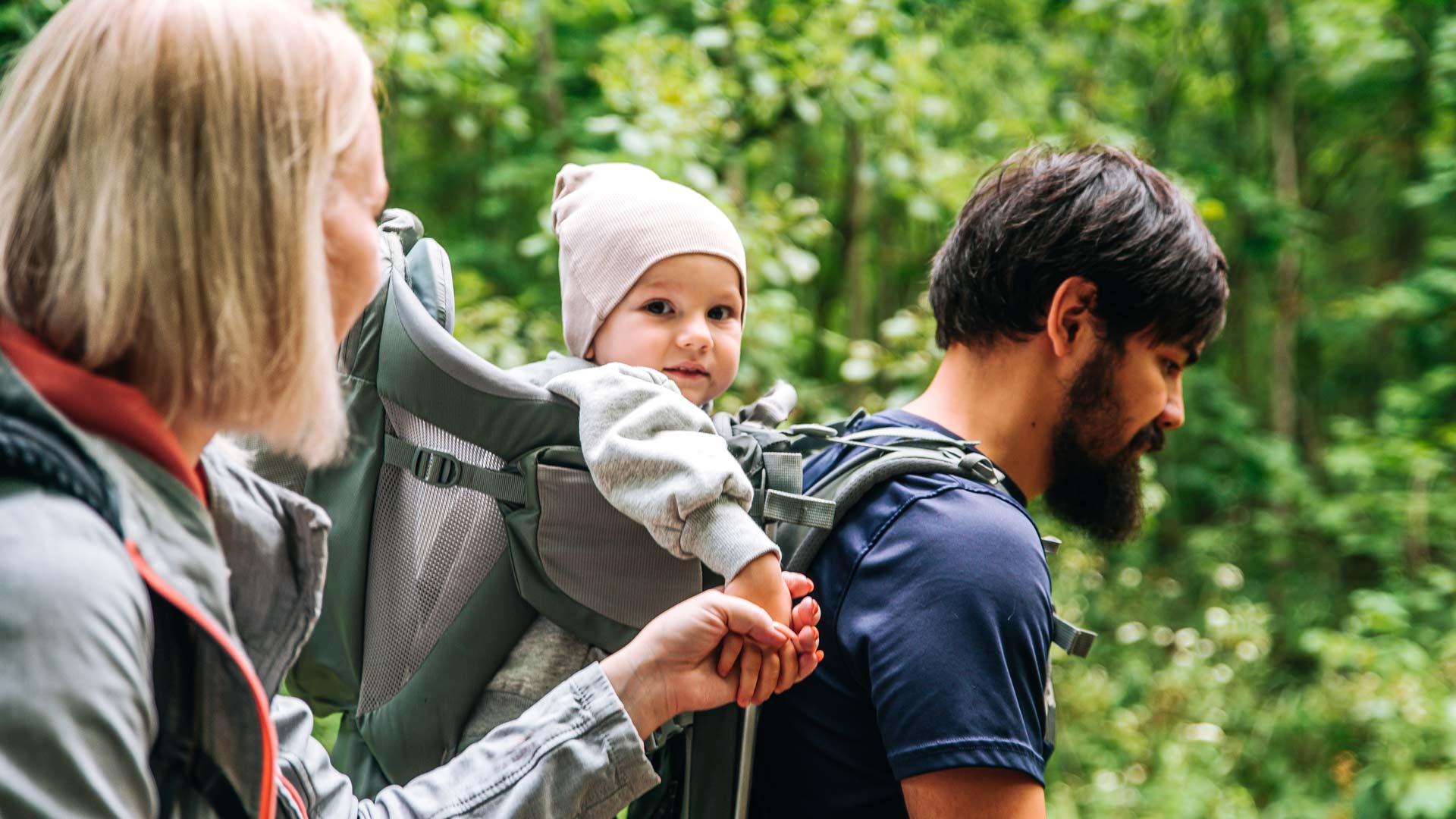 Couple walking through woods with baby in a grey carrier on parents back. 