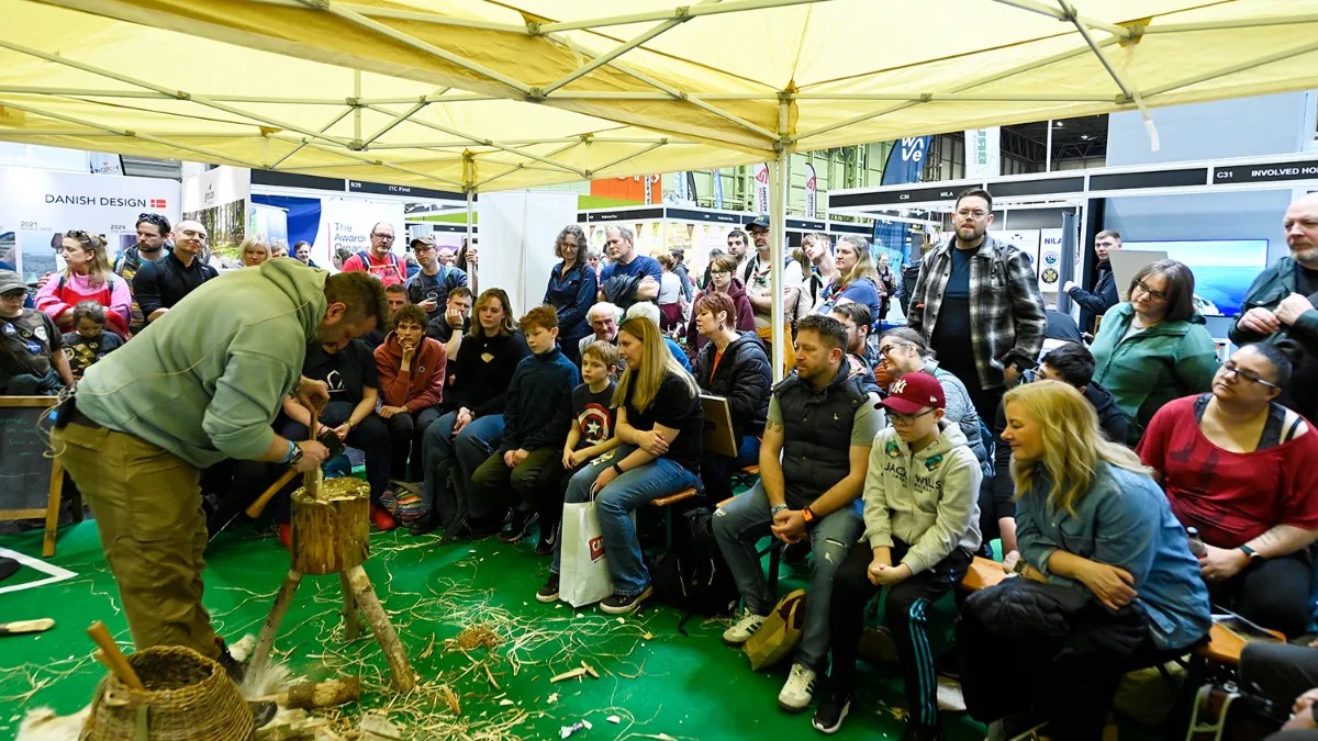 A crowd of people sit under a large yellow canopy watching a woodworking demonstration, as a craftsman carves wood using traditional hand tools.