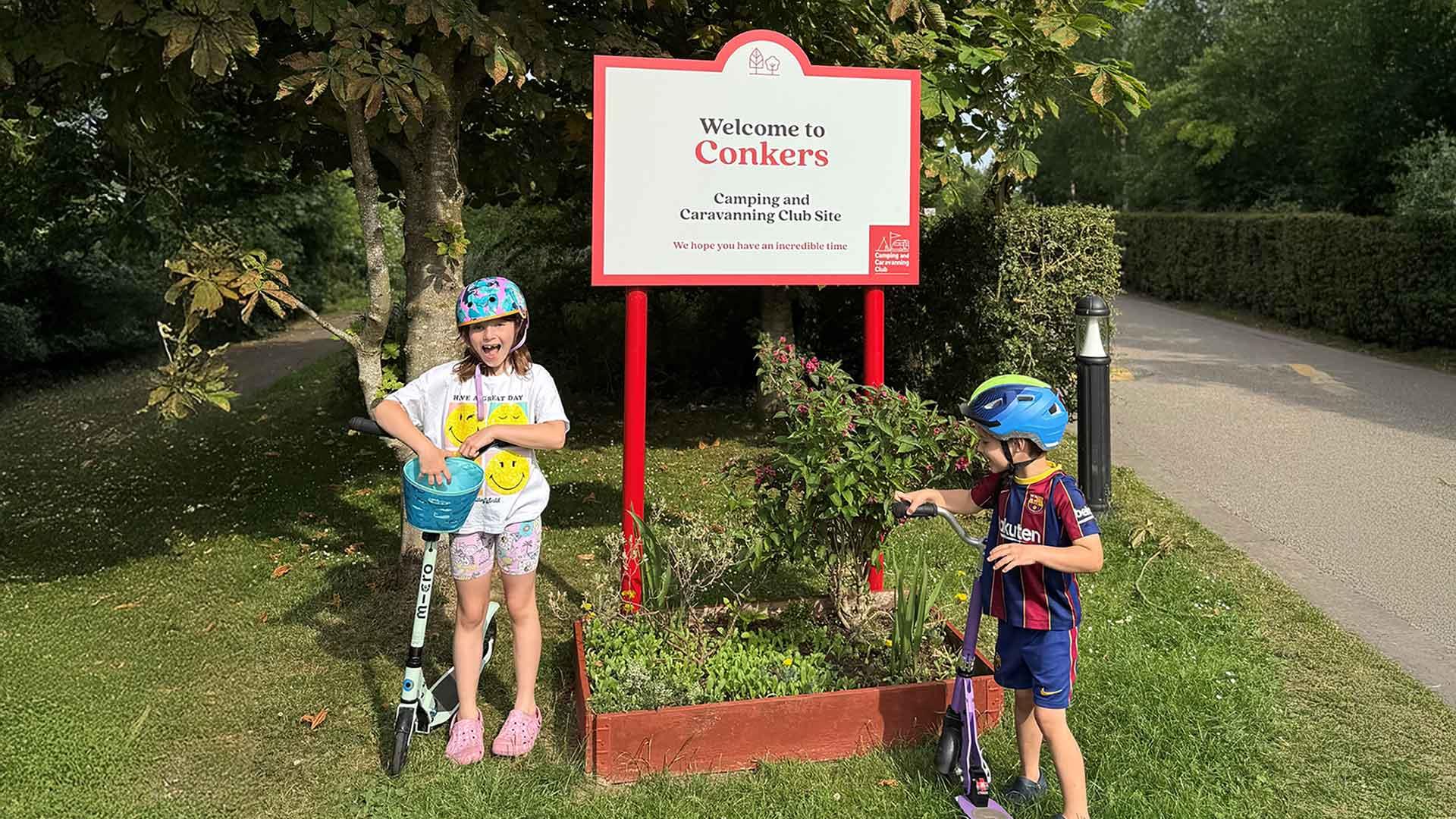 Two children stand with their scooters by the Welcome to Conkers Camping and Caravanning Club site
