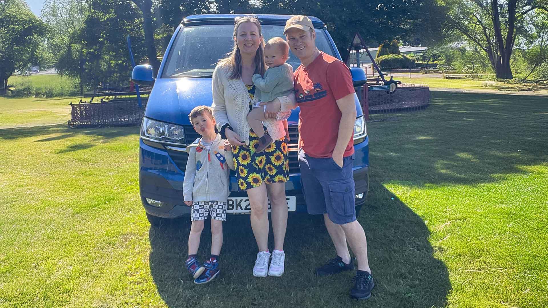 A smiling family of four standing in front of their car on a sunny day looking happy after a fun day out.