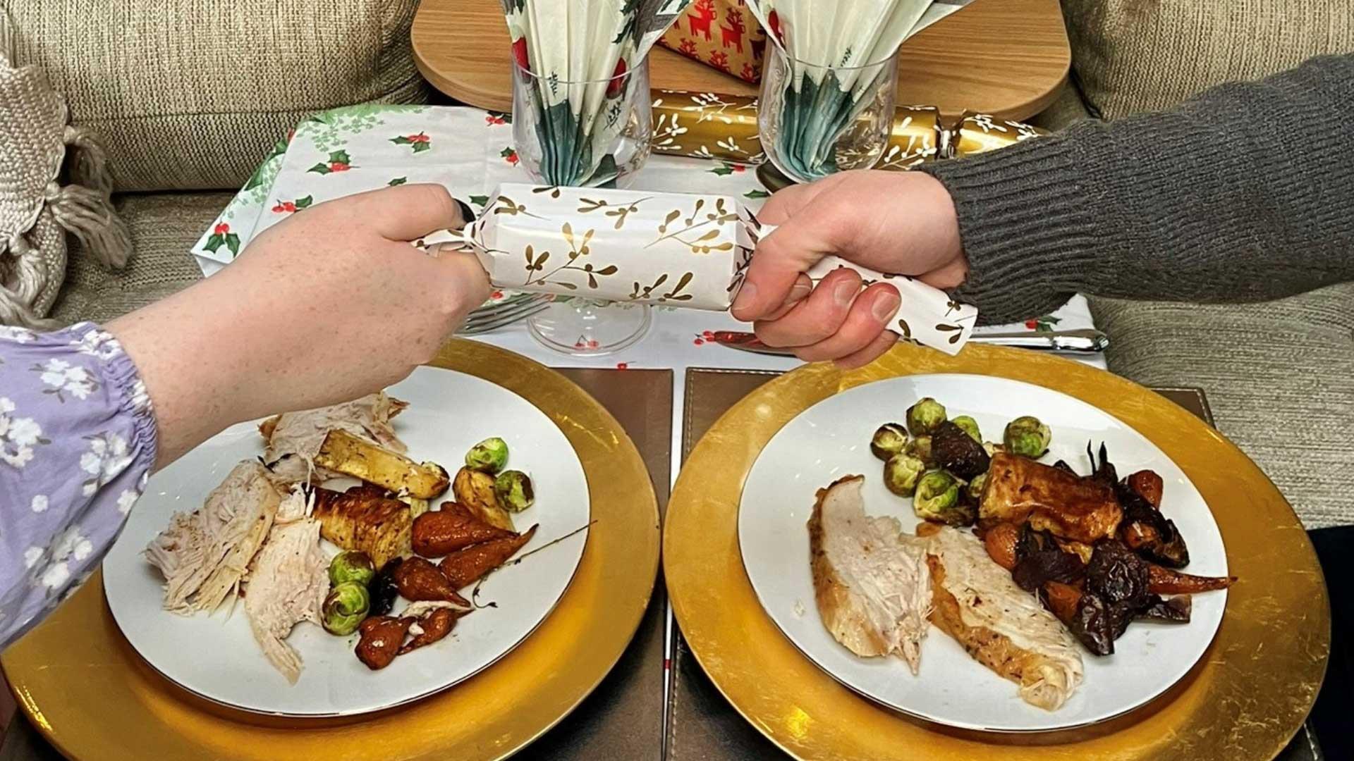 Two hands pull a Christmas cracker over plates of traditional Christmas dinner.