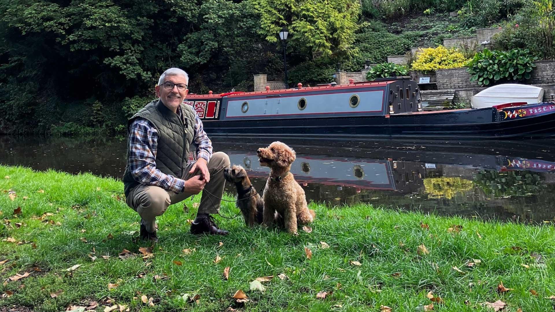 Man kneeling beside two dogs on grassy canal bank with a narrowboat in the background.