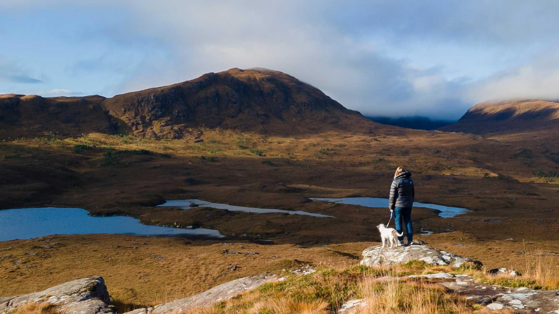 A woman with a dog stands on a low rock, looking out over rugged hills with small pools of water in the foreground.