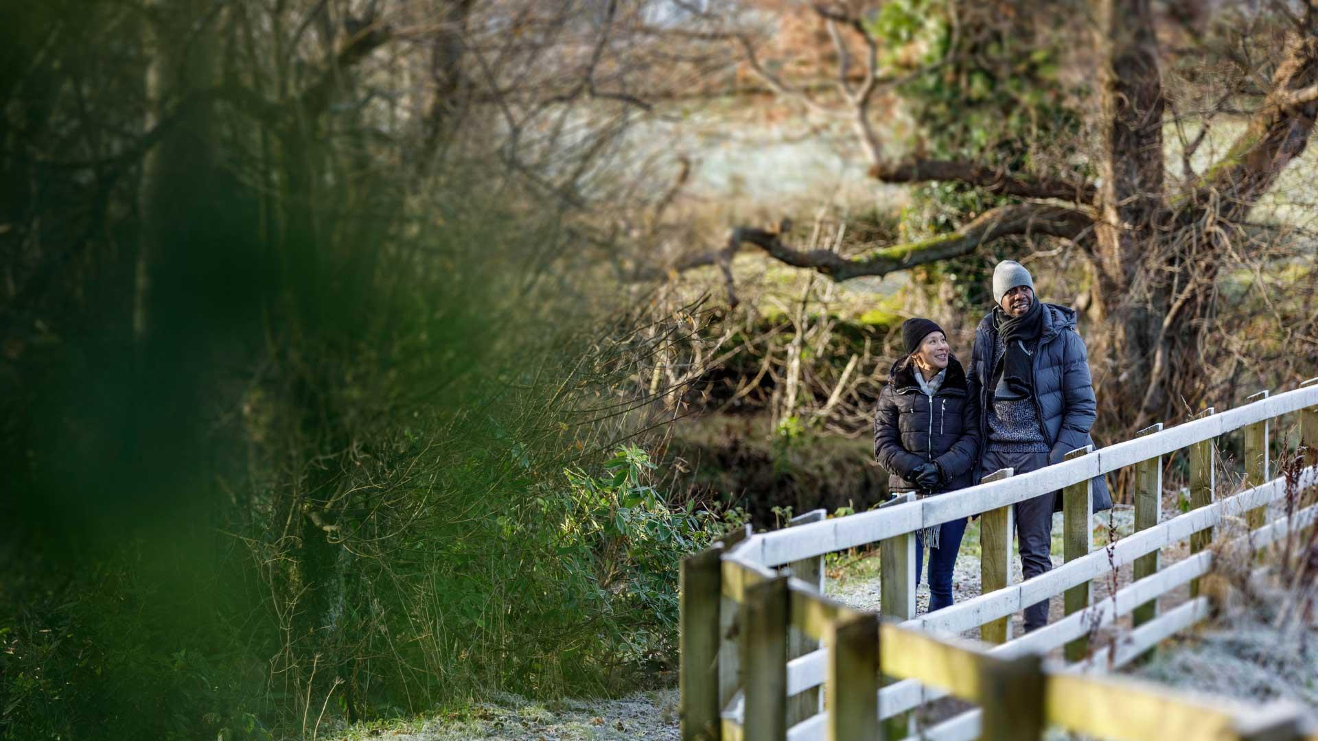 A couple dressed in warm winter clothing walk together along a wooden railing path in a frosty woodland. They are smiling, surrounded by bare trees and soft green foliage in the foreground.