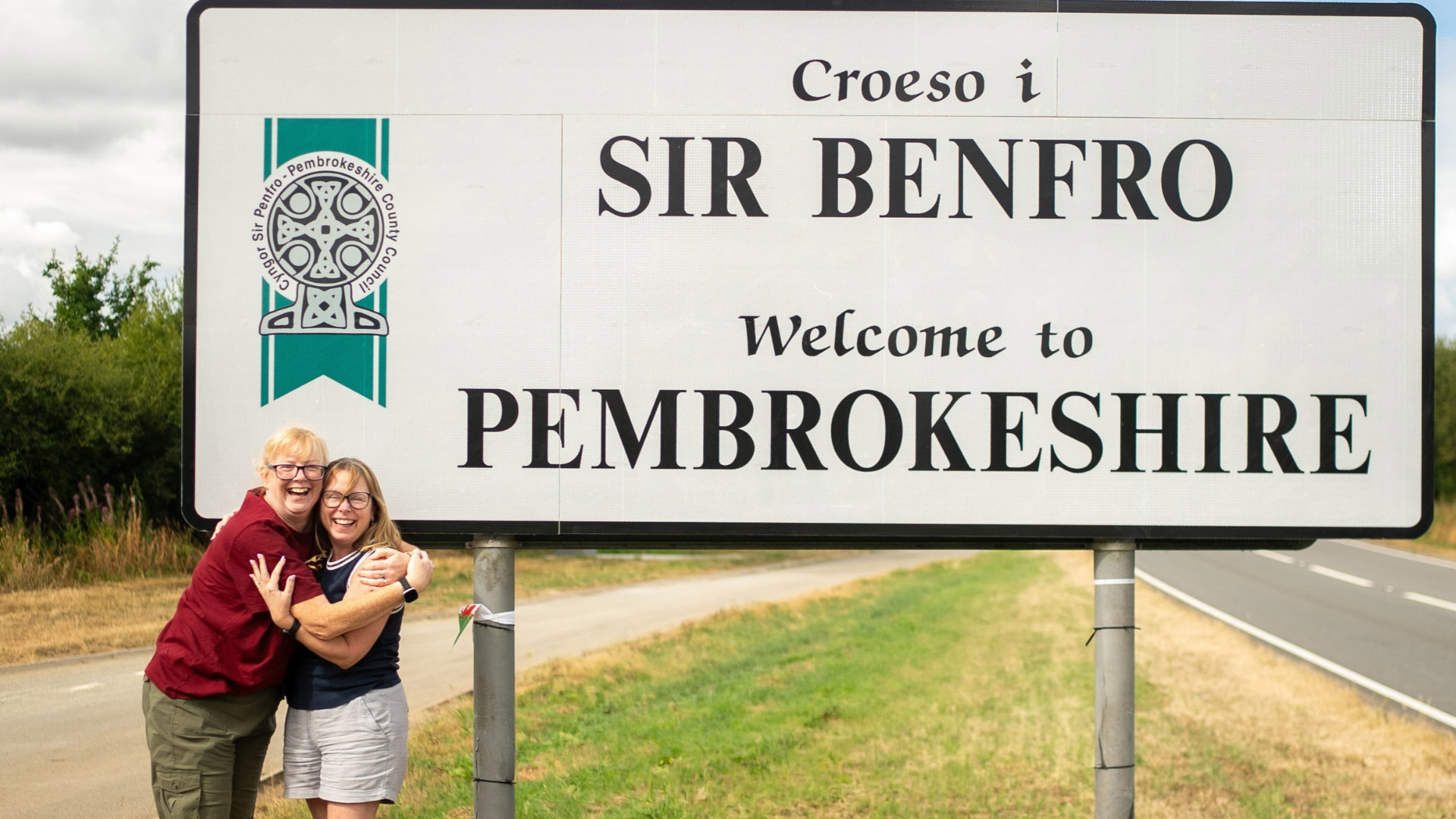 TV travel companions Tricia Sail and Cathie Rowe smiling and hugging in front of the Welcome to Pembrokeshire sign.