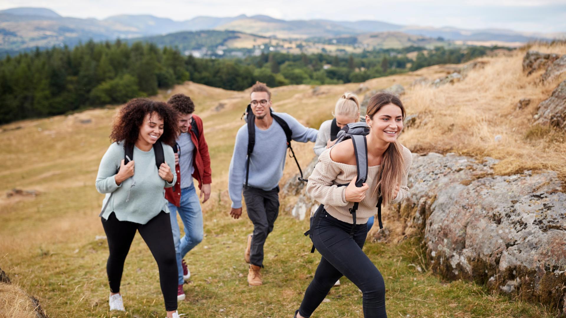 Five young friends smile while hiking up a grassy trail alongside a rocky outcrop. They carry backpacks as they walk, with rolling green hills and mountains visible in the distant background.