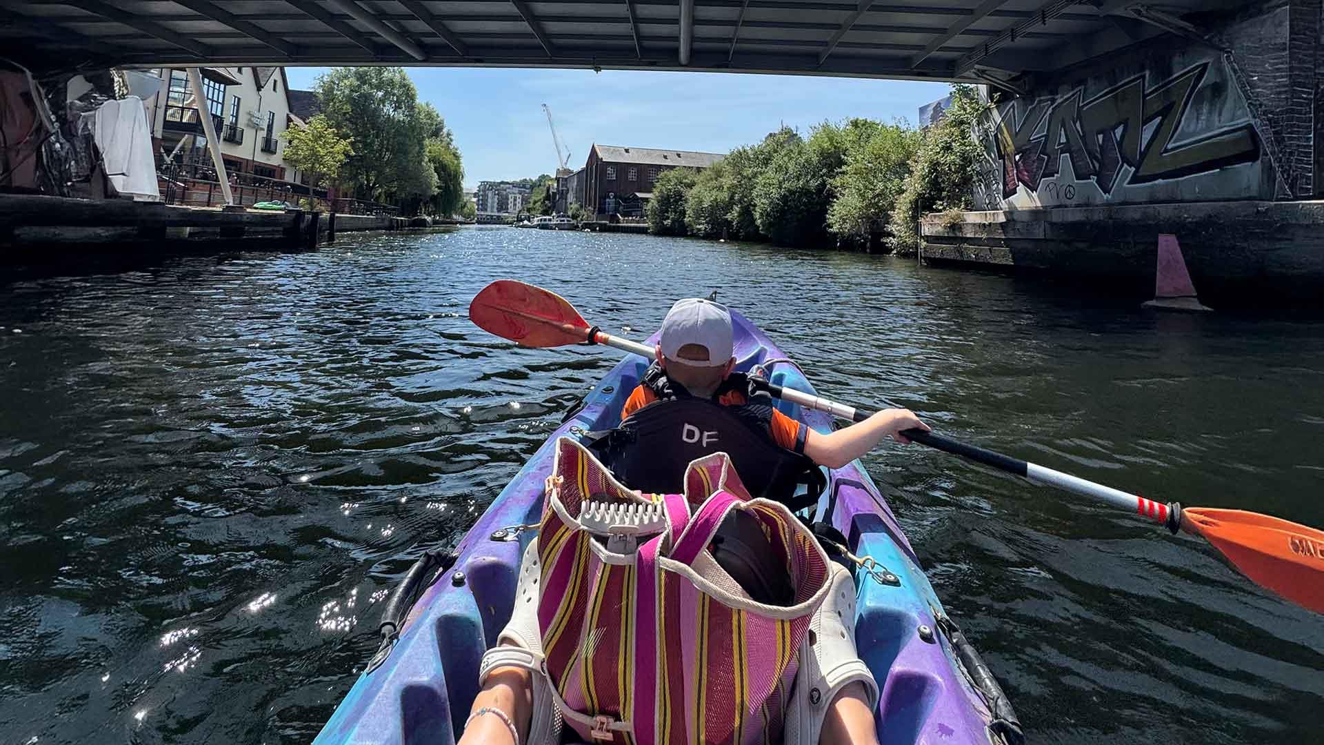 A young boy in a purple kayak rows down a river passing under a bridge