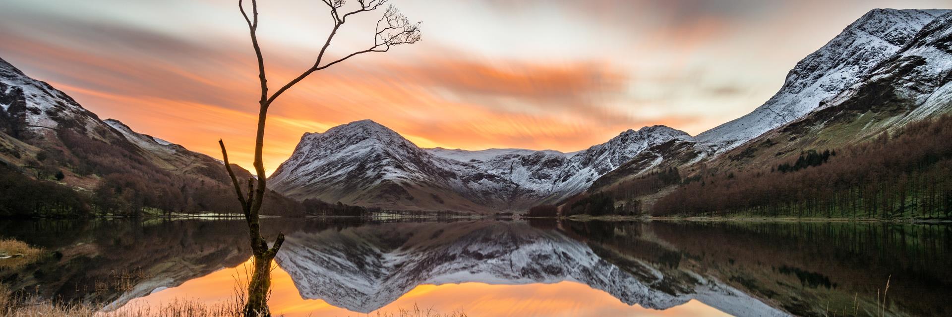 Vibrant sunrise with snowcapped mountains reflecting in calm still water at Buttermere, Lake District, UK